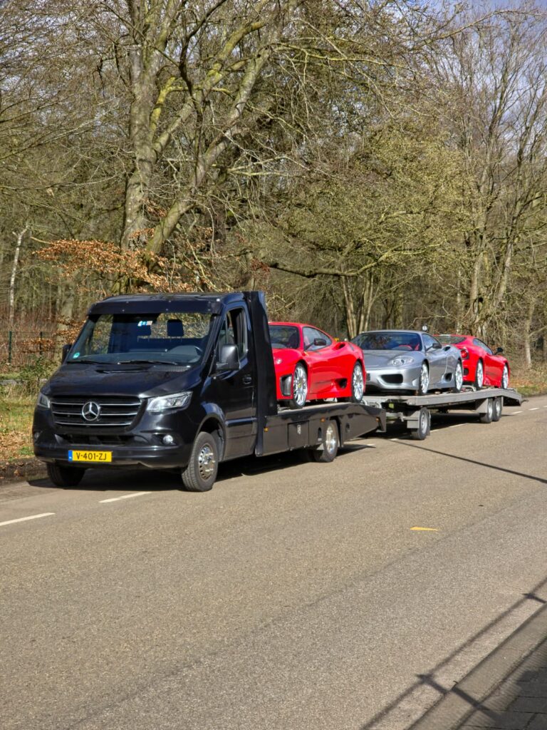 Zwarte Mercedes Sprinter met open oplegger en drie sportwagens onderweg